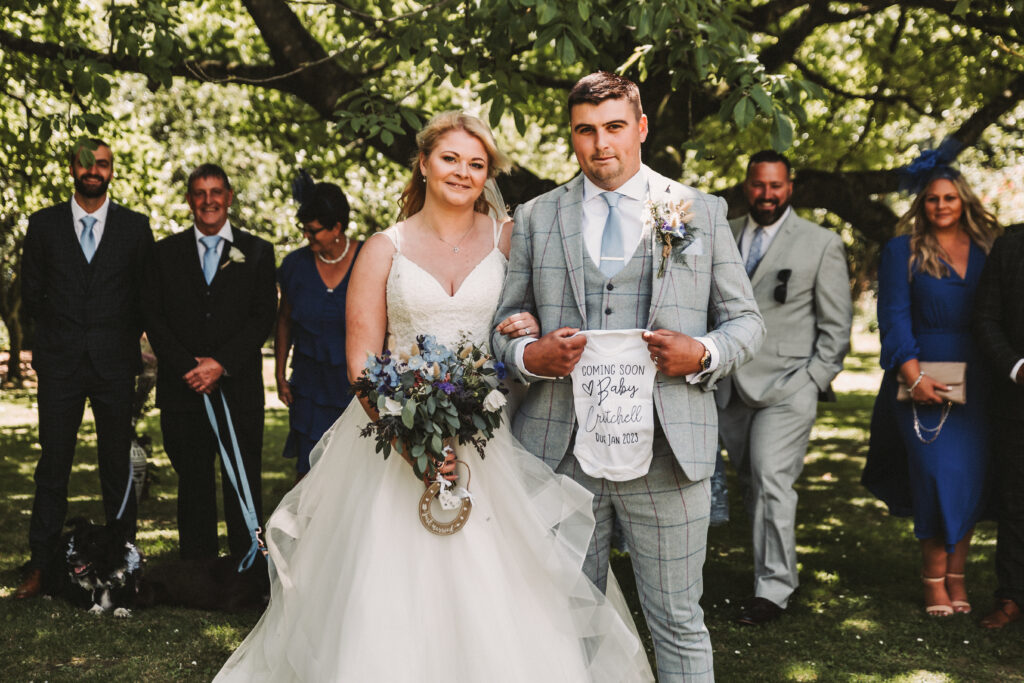 bride and groom standing holding a coming soon baby grow in front of all of their family