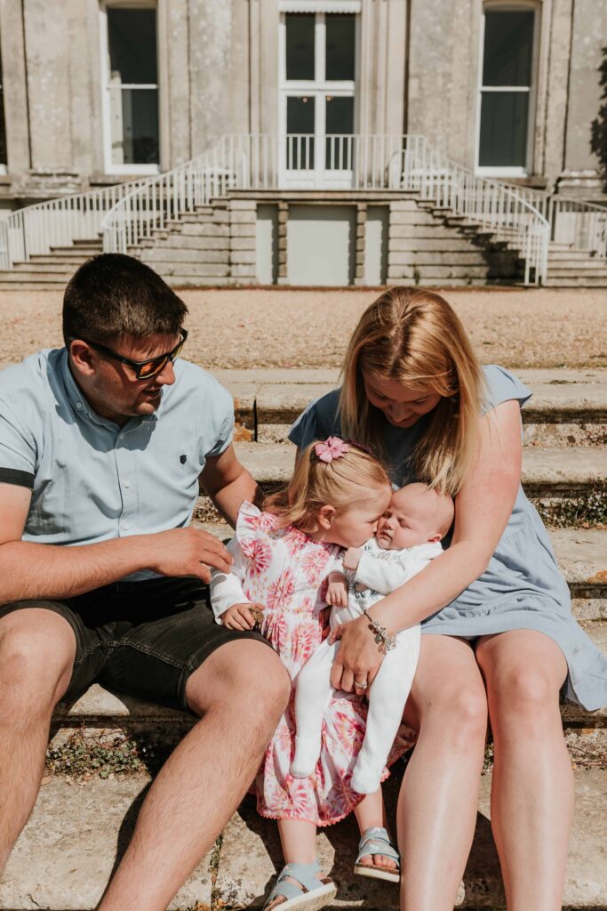family of four sitting on steps whilst little girl kissing her baby brother