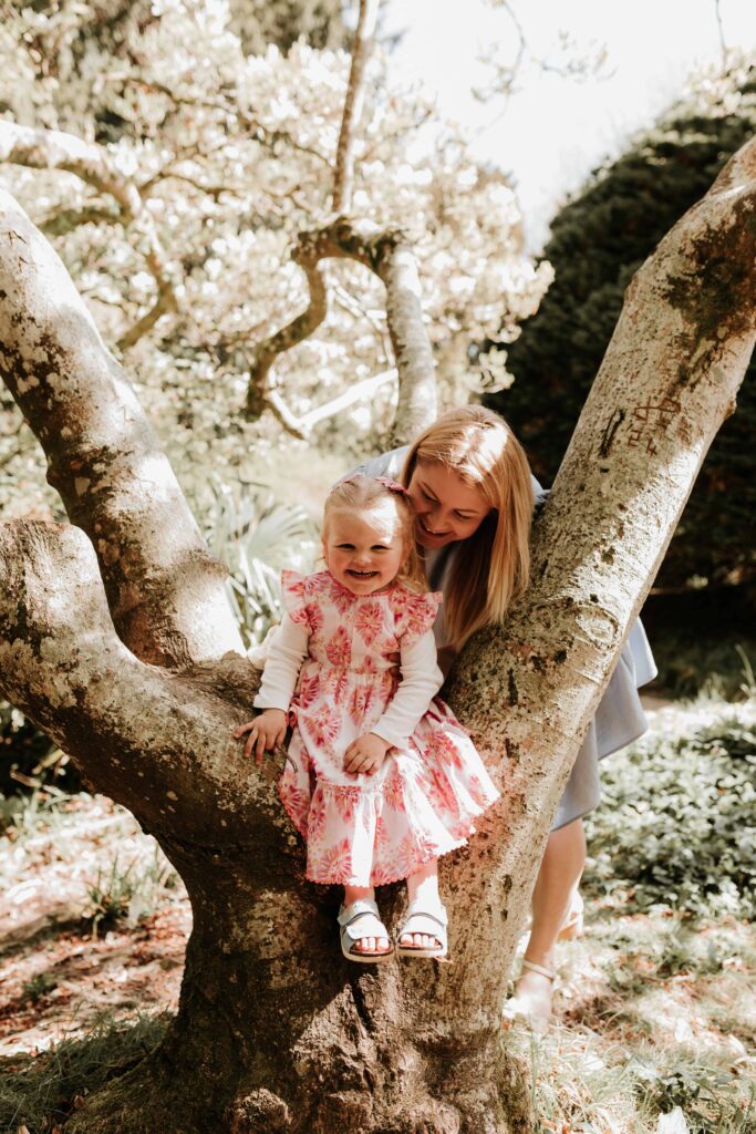 little girl sat in the tree with mum smiling at her from behind