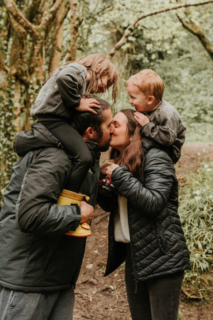 mum and dad kissing with their son and daughter on their shoulders at family photoshoot in cornwall