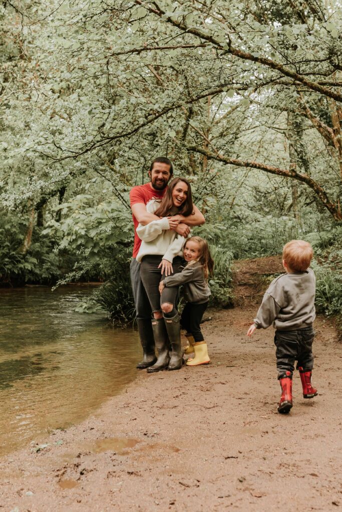 mum and dad standing together near river whilst daughter and son runs towards them
