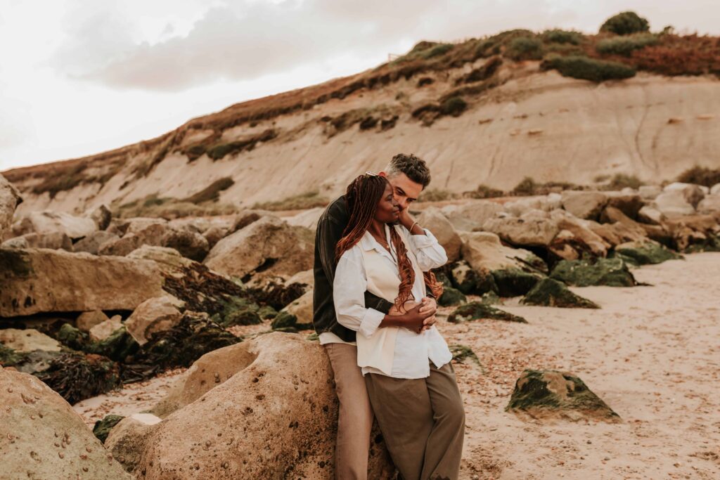 man cuddling woman from behind on a beach leant against a rock