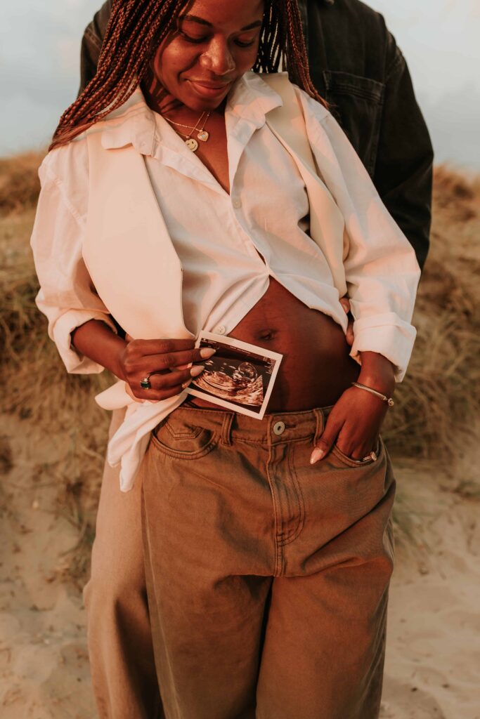 mand stood behind woman whilst she has her belly out holding a baby scan photo