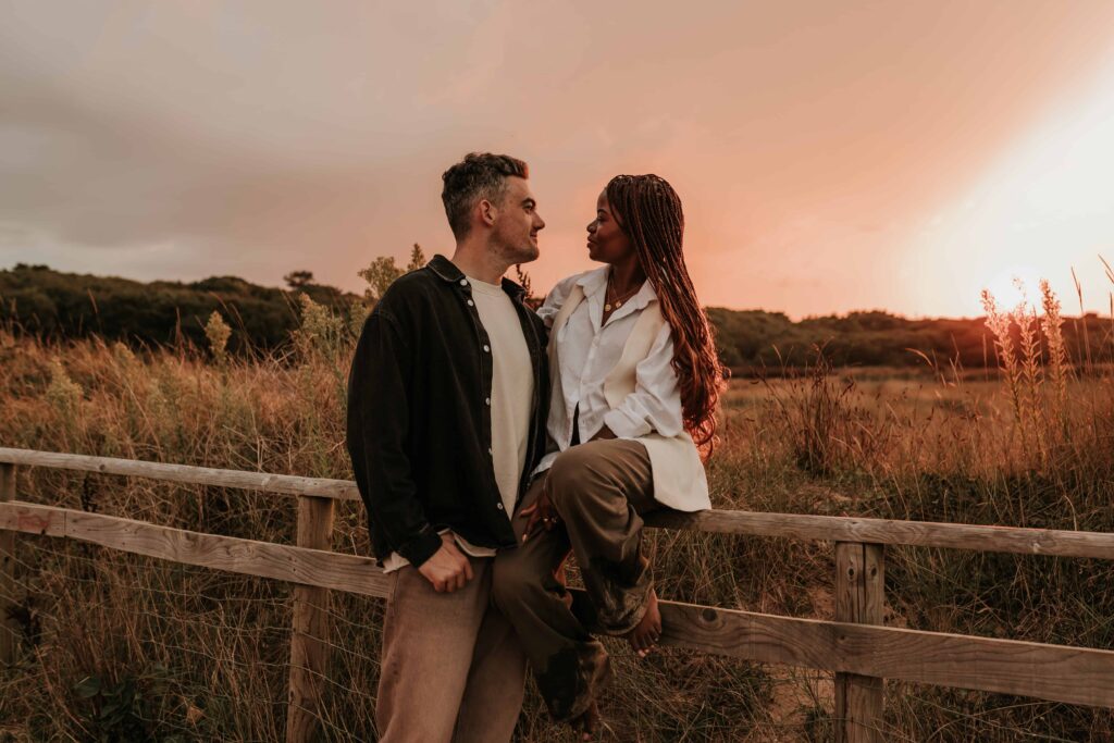 man with arm around woman whilst she sits on a fence during sunset