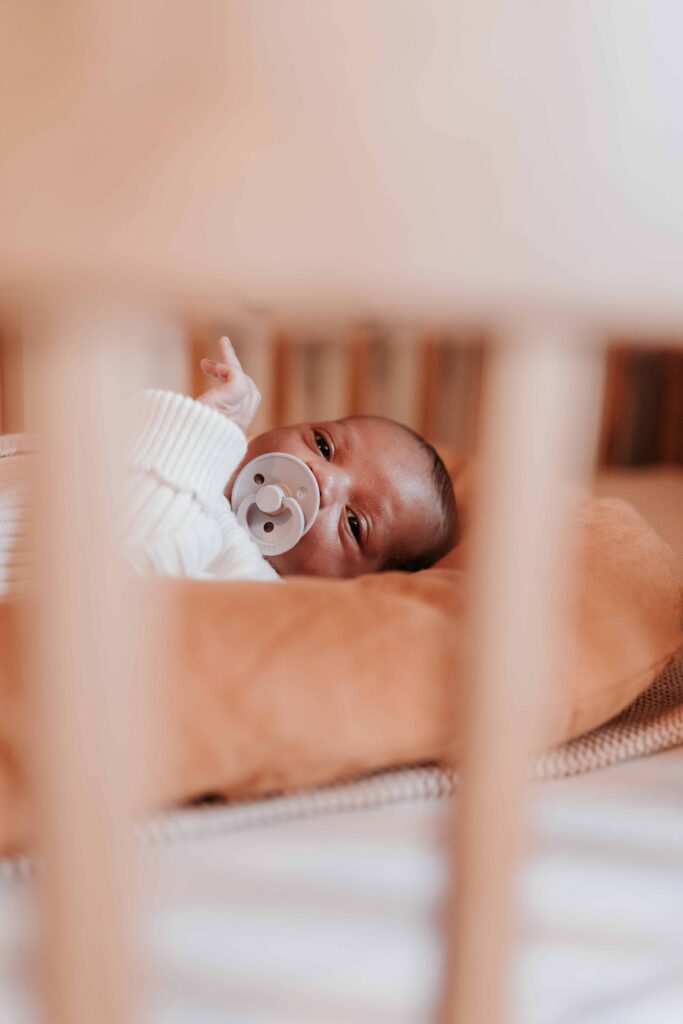 newborn baby with dummy in mouth looking through cot bars at maternity & newborn photoshoot