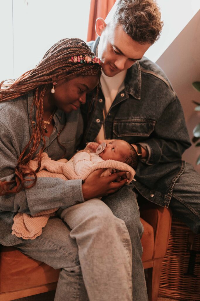 mum holding newborn baby whilst dad sits on the arm of the chair looking over them at maternity & newborn photoshoot