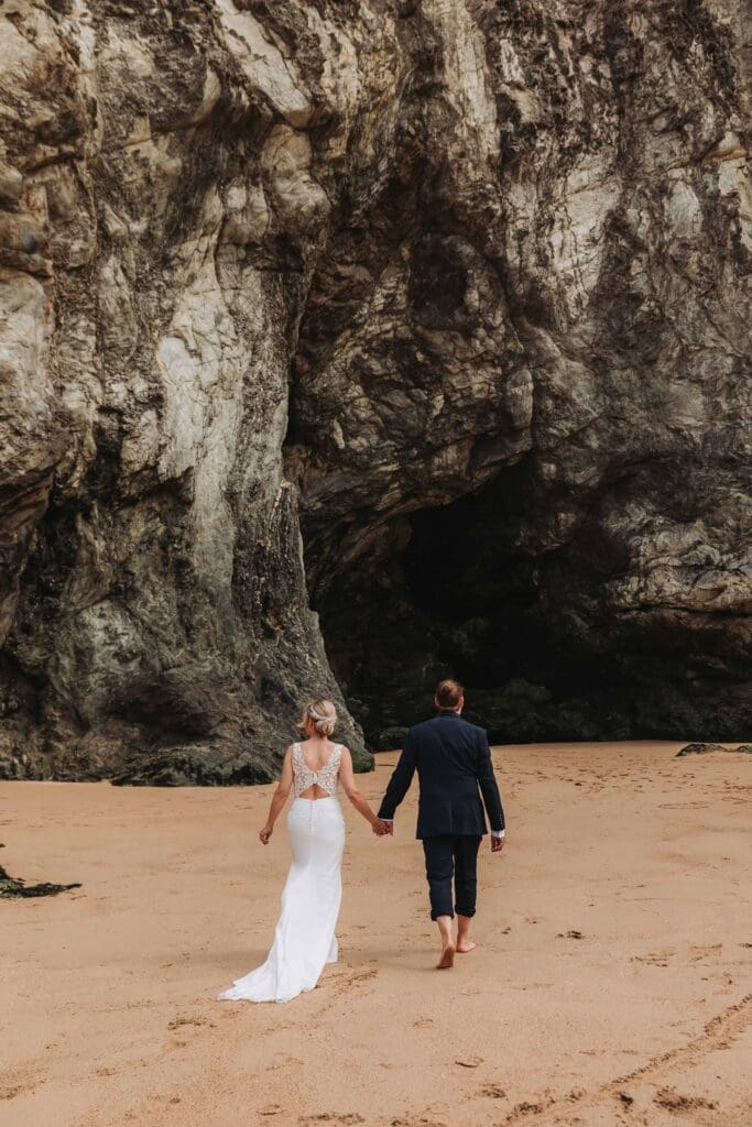 bride and groom walking towards rock face at holywell bay